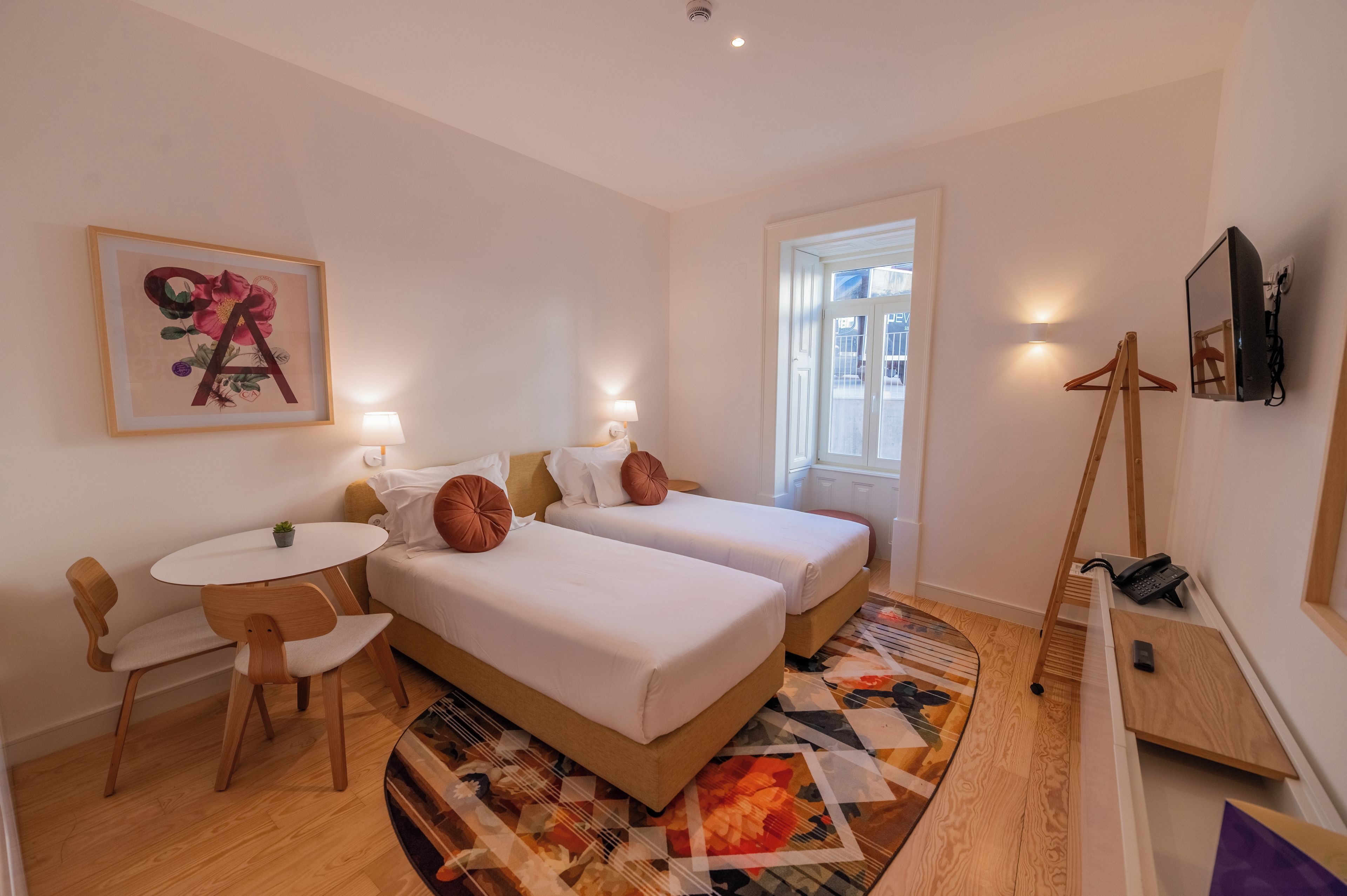 A hotel room with two beds, a small table, a chair, a desk, a desk lamp, a mirror, a coat rack, and a window.