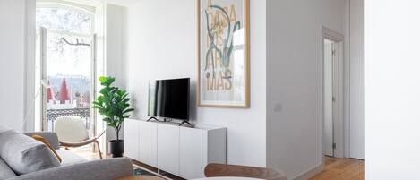 A modern living room with a grey sofa, a round table with a potted plant, a flat-screen TV on a white cabinet, and a large framed artwork on the wall.