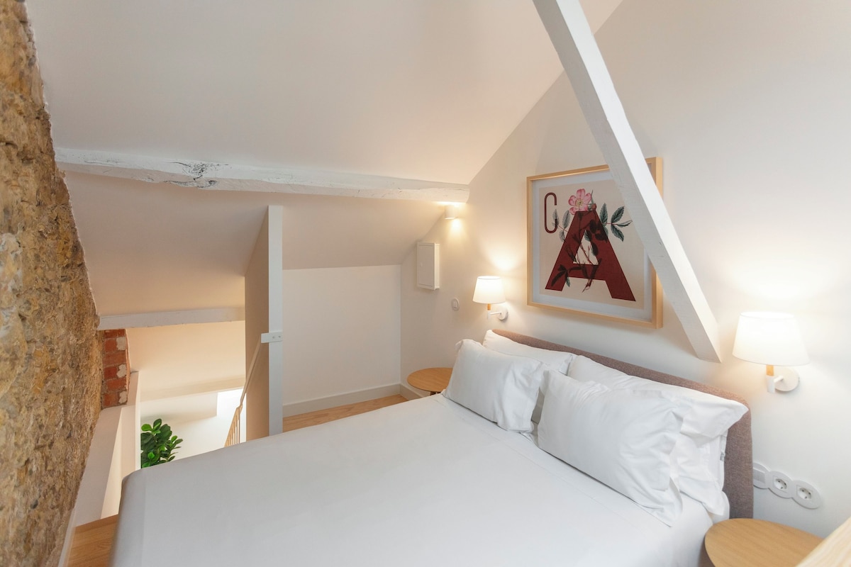 Une chambre à coucher avec un lit, deux lampes de chevet, un tableau accroché au mur et une prise électrique visible.