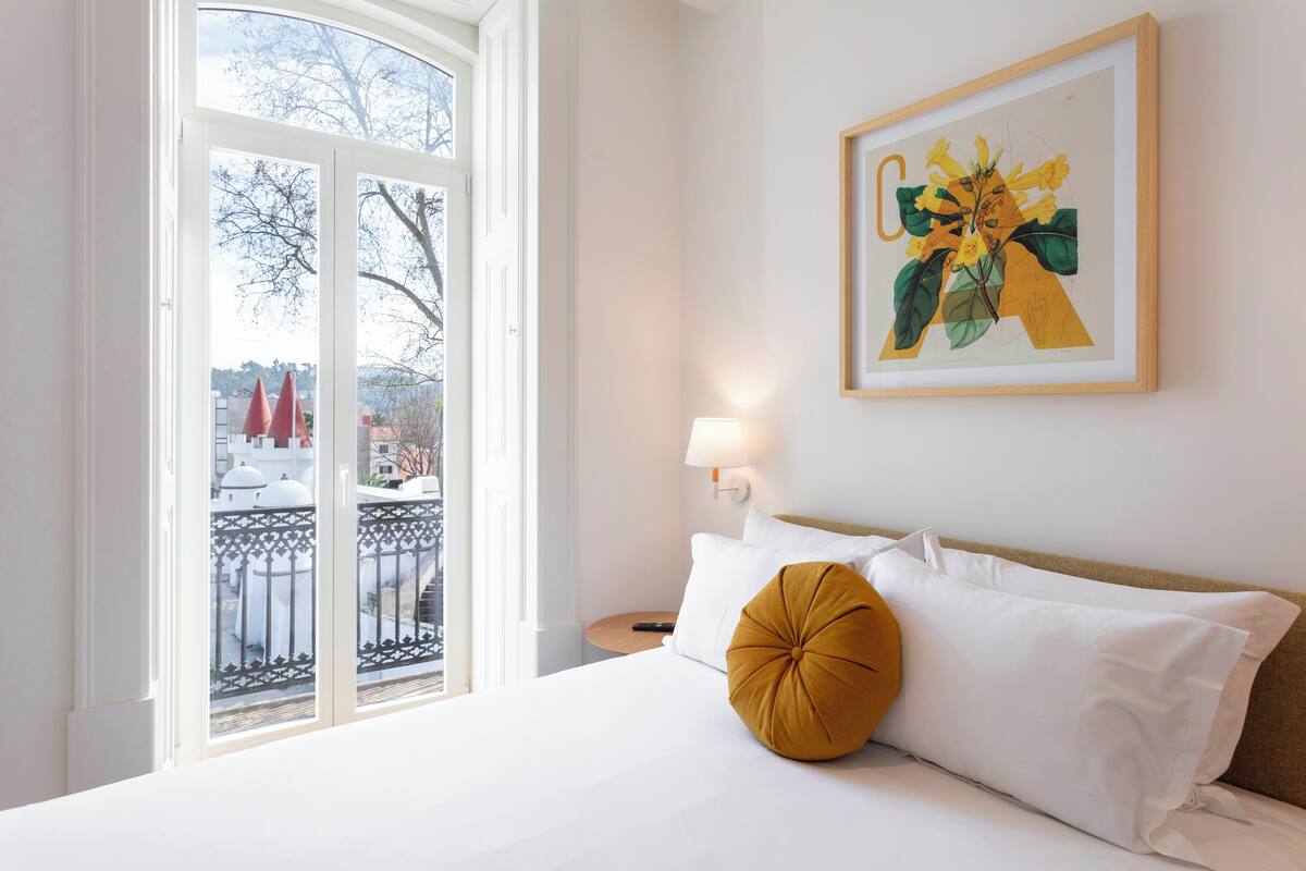 A hotel room with a bed, a window with a view, a framed artwork on the wall, and a bedside table with a lamp.