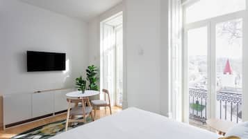 Quarto de hotel com cama, televisão fixada na parede, pequena mesa de jantar com cadeiras, planta em vaso e varanda com vista para a cidade.