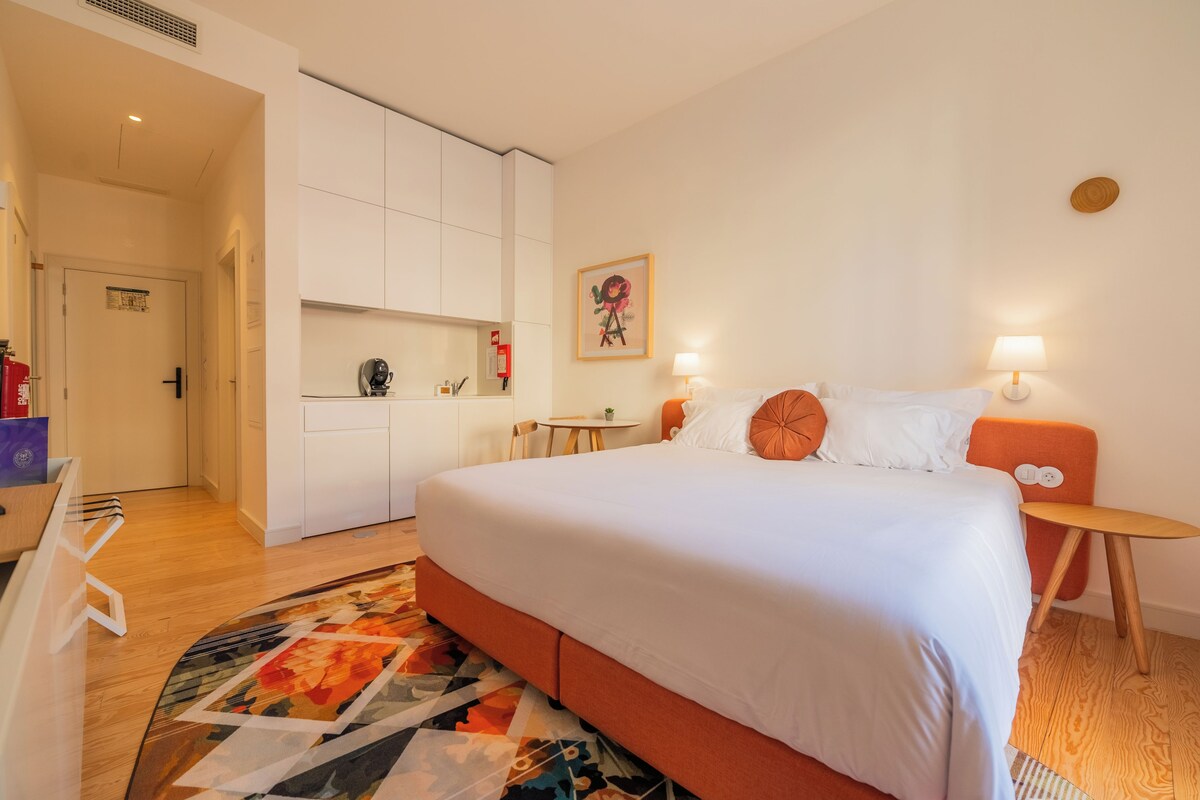 A hotel room with a bed, bedside tables, a small table, a chair, a rug, a wardrobe, and a door.