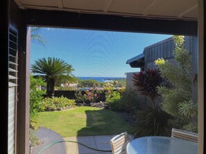 A view through a window with a garden, a palm tree, and a building with a flat roof. - Secluded , cozy poolside getaway.. Ocean views, near black sand beach & turtles. (Pahala)