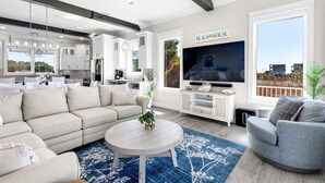 Ein modernes Wohnzimmer mit Sofa, Sessel, Fernseher und einer Küche im Hintergrund.