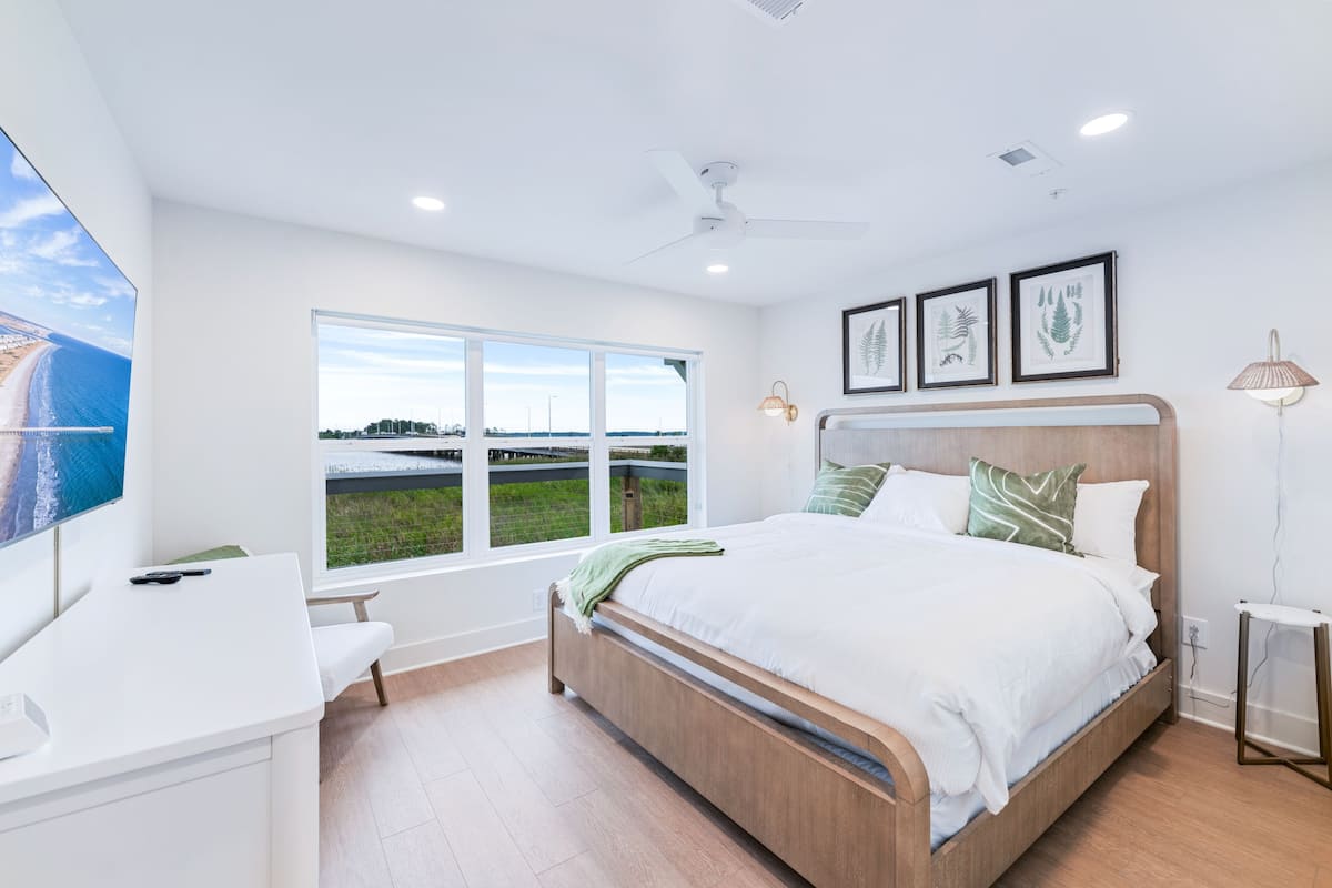 Un dormitorio moderno con una cama grande, un escritorio y una ventana que ofrece vistas a la playa y al paisaje urbano.