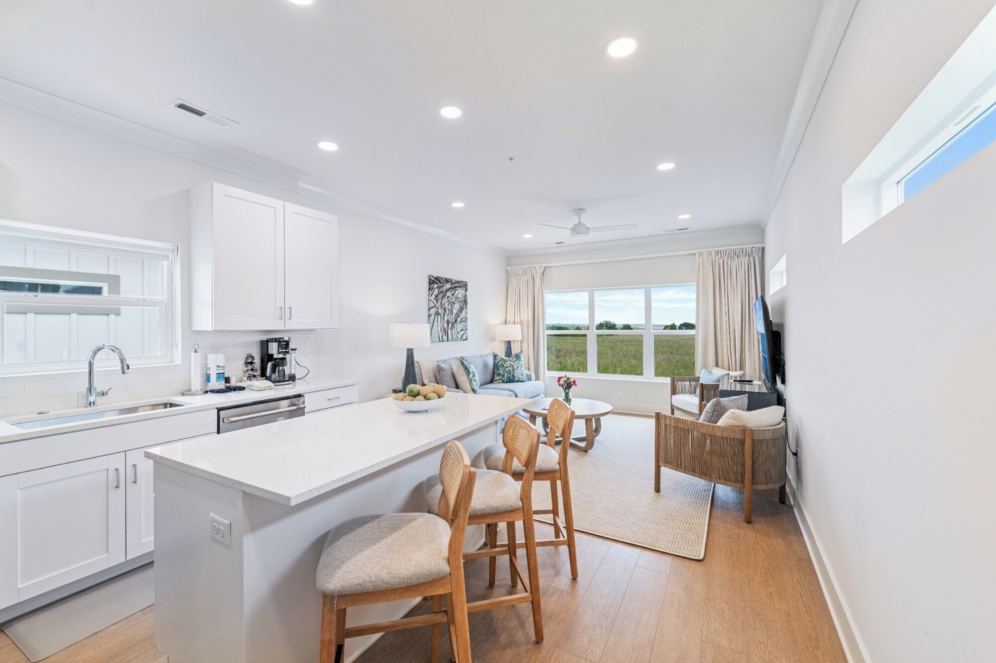 A modern kitchen with white cabinetry, a large island with bar stools, a dining area with a table and chairs, and a large window with a view of the outdoors.