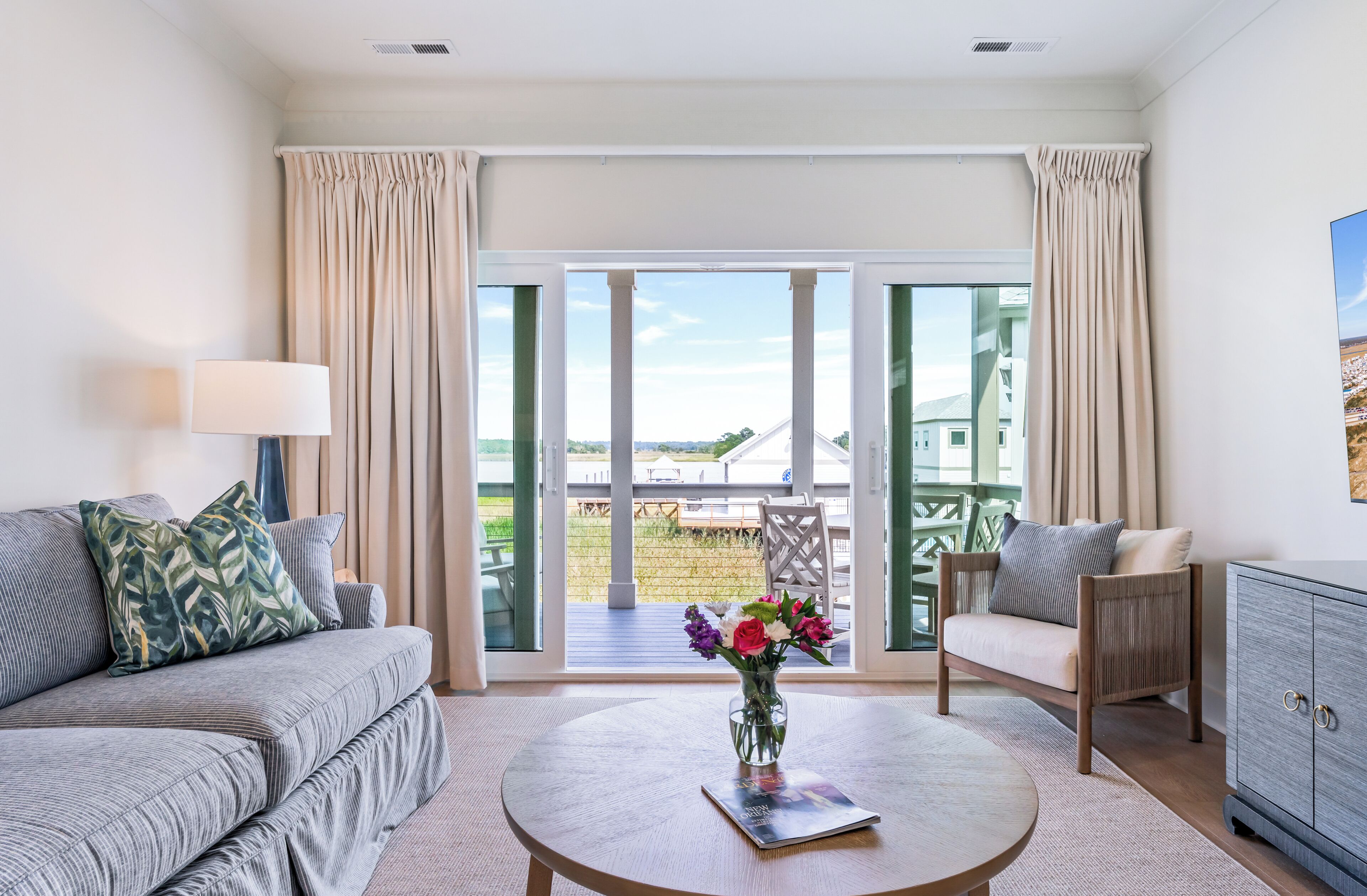 A modern living room with a sofa, a coffee table with a vase of flowers, a side table with a lamp, and a view of the outdoors through large sliding glass doors.