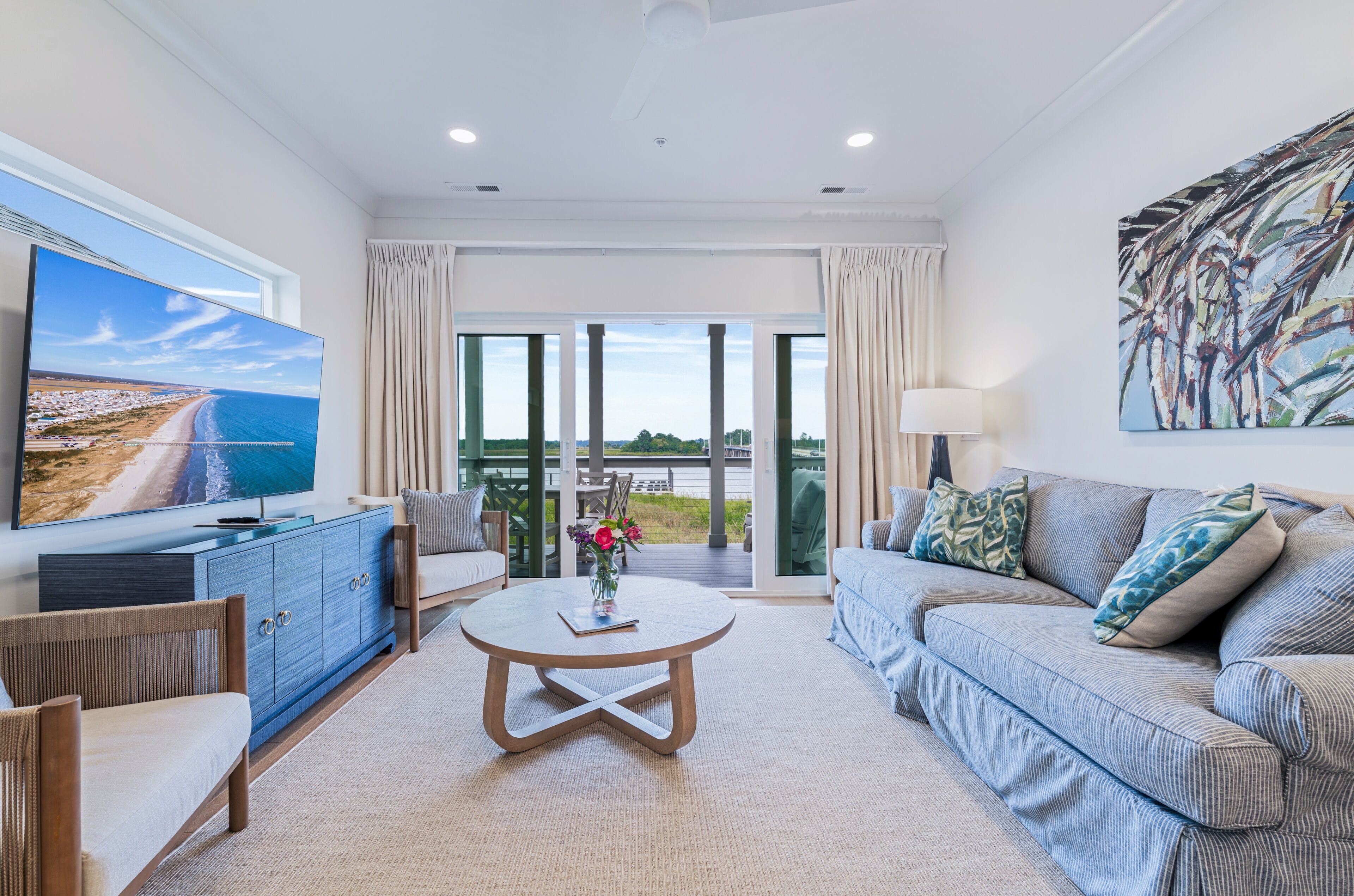 A modern living room with a flat-screen TV, a round coffee table, a sofa, and a large window with a view of the sea.