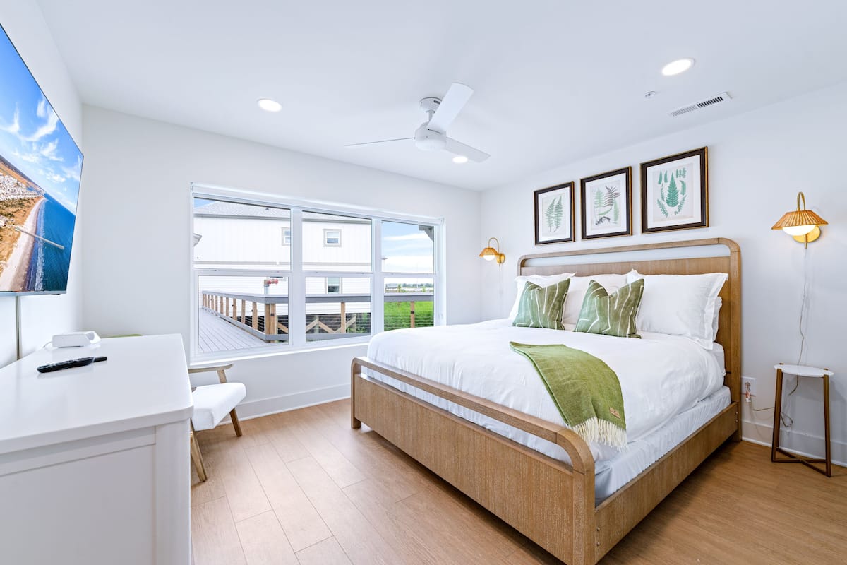 Un dormitorio con cama, escritorio, silla, ventana, ventilador de techo y cuadros en la pared.