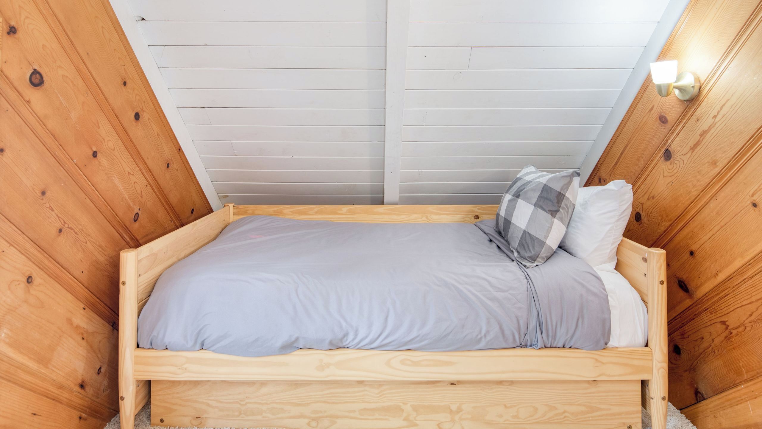 A wooden-framed bed with a grey mattress and checkered pillow in a room with sloped wooden walls and a skylight.