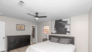 Une chambre à coucher comprenant un lit, une commode, un ventilateur de plafond, des œuvres d’art et une fenêtre avec des stores.
