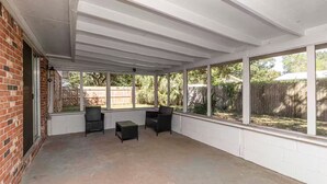 Une terrasse couverte avec un mur en briques, deux fauteuils en osier, une petite table et de grandes fenêtres donnant sur une cour arrière avec une clôture en bois et des arbres.