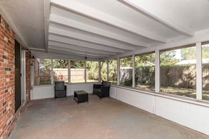 Un patio techado con un muro de ladrillo, dos sillas de mimbre, una mesita y ventanas amplias que dan a un jardín trasero con cerramiento de madera y árboles.