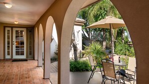 A covered patio with a table and chairs, a door with glass panels, and a view of greenery.