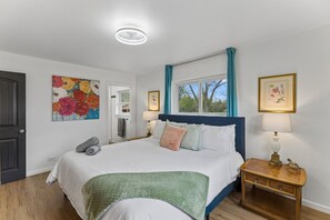 Une chambre à coucher avec un lit, une table de chevet, une lampe, une fenêtre avec des rideaux, une porte et un tableau au mur.
