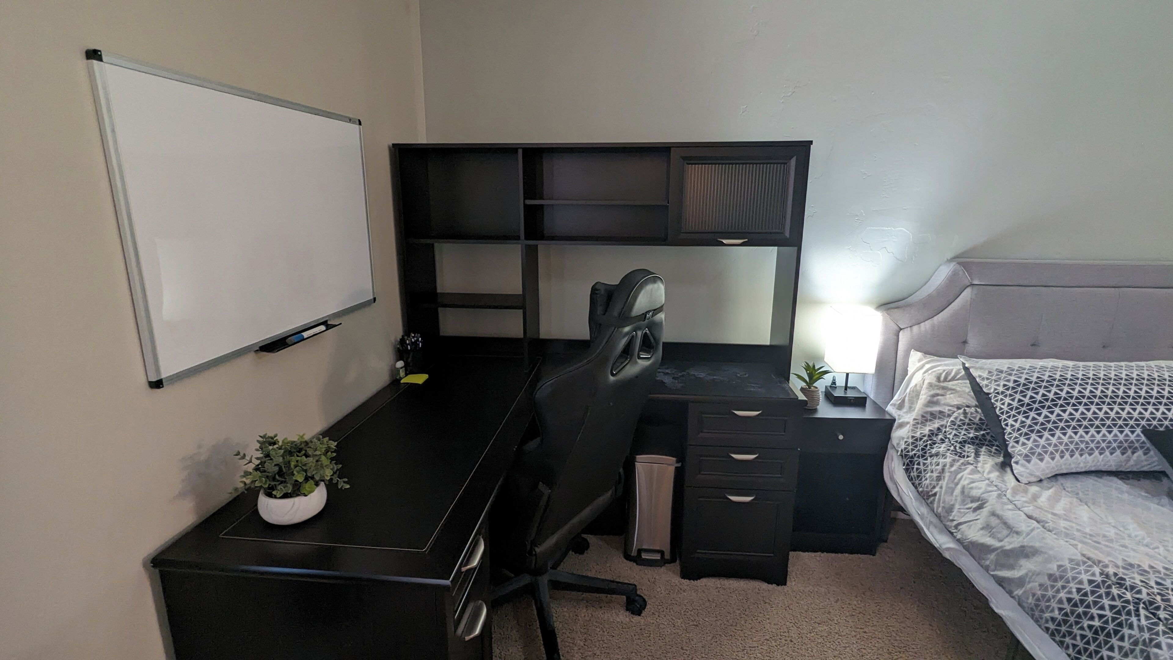 Une chambre à coucher avec un lit, un bureau avec une chaise, un tableau blanc et une petite plante.