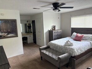 Ein Schlafzimmer mit einem Bett, einem Schminktischchen, einem Kleiderschrank, einem Spiegel, einem Deckenventilator und einem Bild an der Wand.