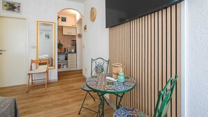 Een compacte leefruimte met een kitchenette, een eethoek met een tafel met dessin, en een aan de muur bevestigde televisie.