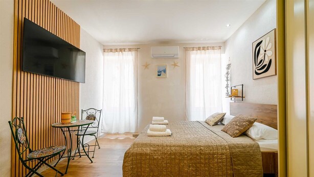 Een slaapkamer met een bed, een aan de muur bevestigde televisie, een klein tafeltje met stoelen en een raam met gordijnen.