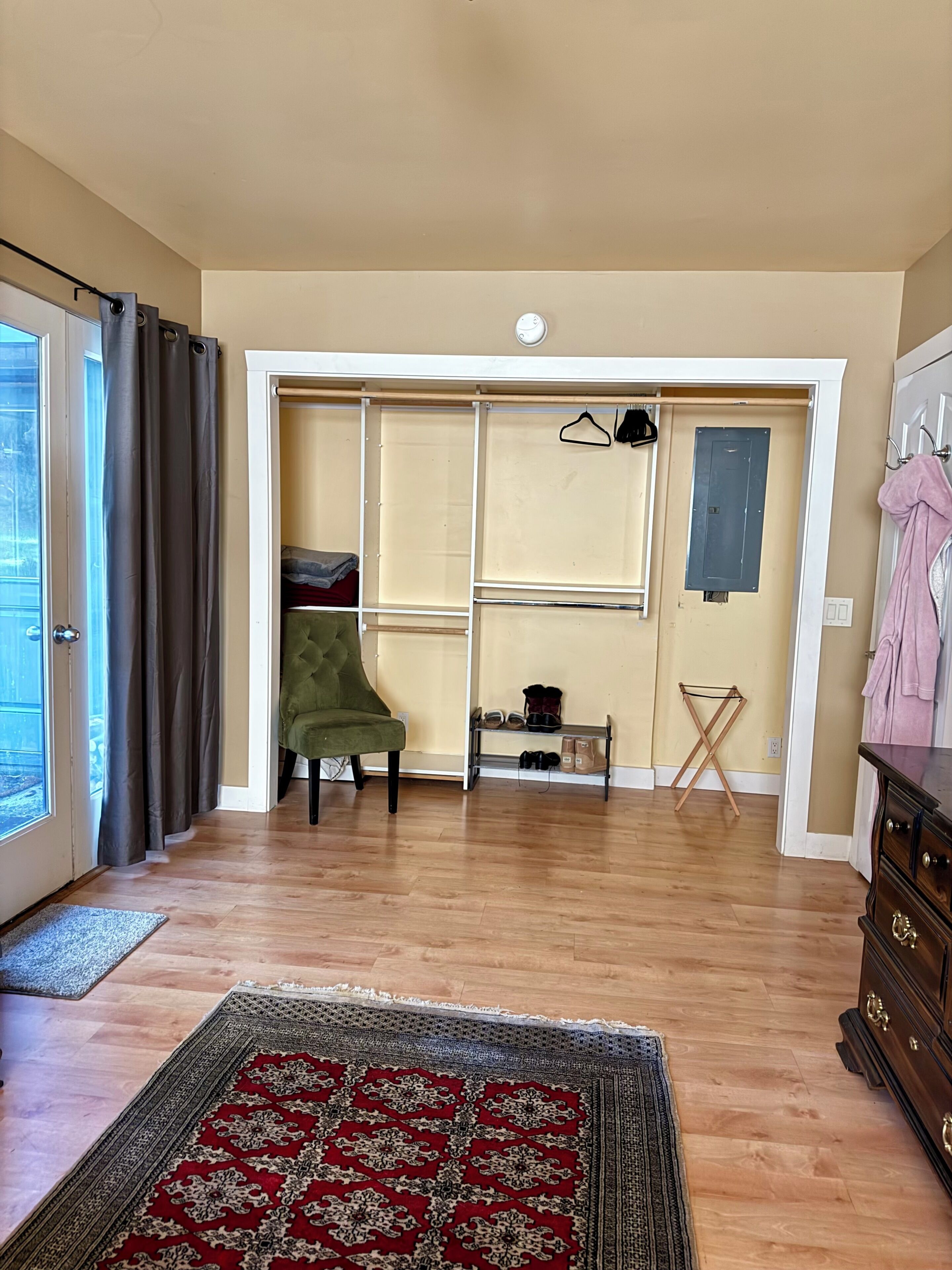 A room with a wooden floor, a green armchair, a patterned rug, a shoe rack, and a dresser.