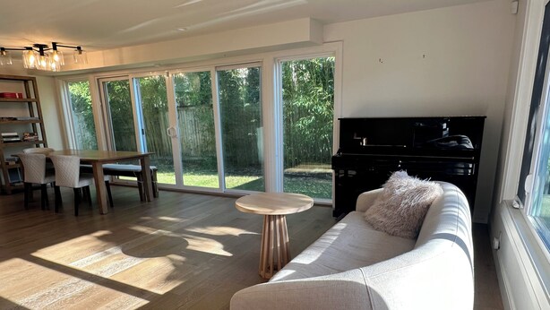 A spacious living room with a sofa, a dining table with chairs, a piano, and large windows with a view of greenery.