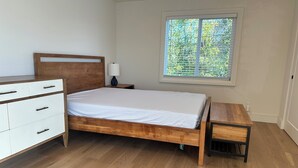 A bedroom with a wooden bed, a white dresser, a bedside table with a lamp, and a window with blinds.