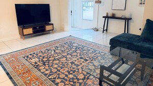 Una sala de estar con televisor de pantalla plana, un sofá oscuro, una mesa de centro de vidrio, un tapete estampado, una consola decorativa y una puerta que da al exterior.