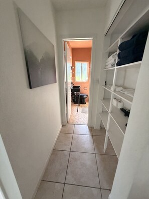A narrow hallway with a tiled floor leading to a bathroom with a toilet and sink. There is a white shelf unit with folded towels and a black bag on the right. - Fort Lauderdale Gem Private Yard and Patio (Fort Lauderdale)