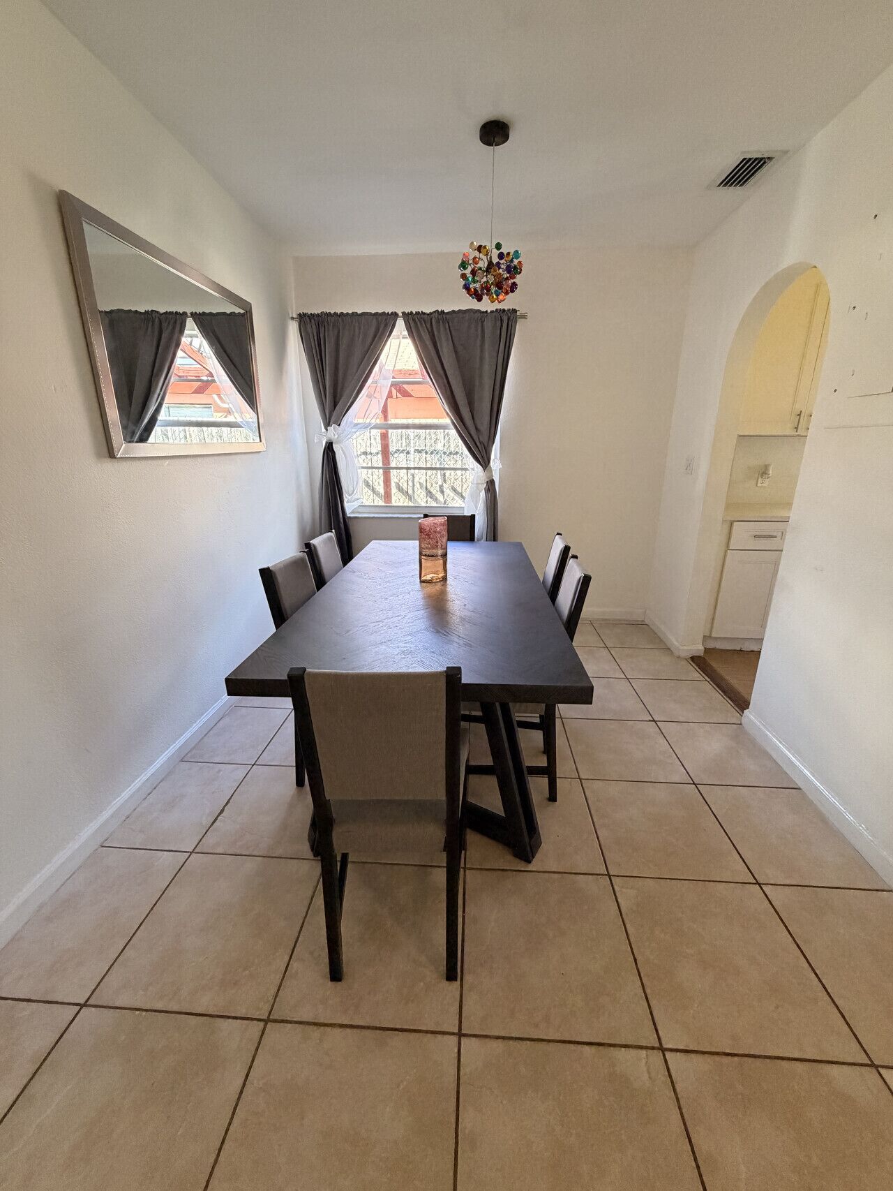 Comedor con mesa rectangular, seis sillas, una araña y vistas a través de una ventana con cortinas.