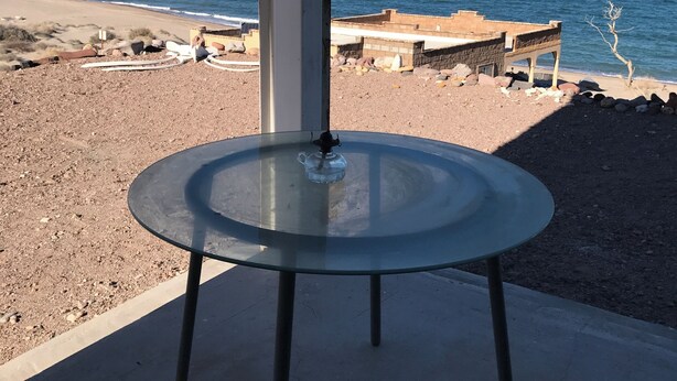 A glass table with a teapot on a concrete floor, overlooking a beach with a clear sky.