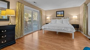 Un dormitorio con cama, cómoda, dos lámparas de noche, un cuadro y una puerta corrediza de vidrio que da a un espacio exterior.
