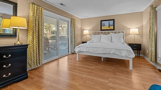 A bedroom with a bed, a dresser, two bedside lamps, a painting, and a sliding glass door leading to an outdoor area. - Windmere Cottage in Charming Pinehurst (Pinehurst)