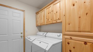 A laundry room with two white front-loading washers and dryers, wooden cabinets above, and a white door. - Pine Cottage (Big Bear Lake)