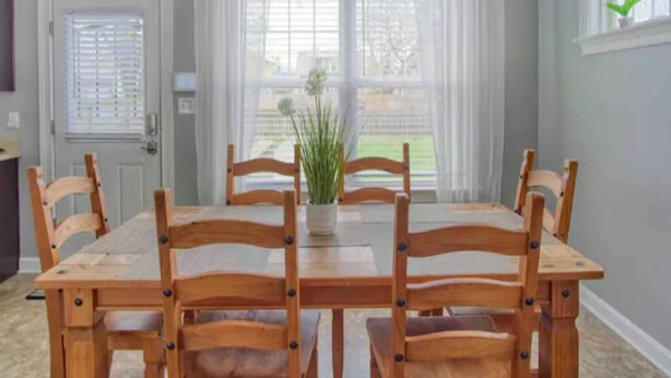 Un comedor con una mesa rectangular y seis sillas de madera, una planta en maceta y vistas amplias a través de grandes ventanales.
