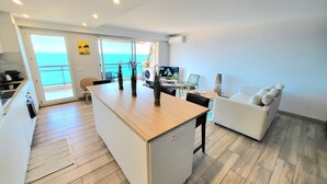 Eine moderne Küche mit Kochinsel, ein Essbereich mit weißem Sofa und Meerblick durch große Fenster.