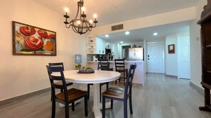 A dining area with a round table and chairs, a chandelier, and a kitchen in the background. - Gorgeous Third-Level Condo Princesa D-308 (Puerto Peñasco)