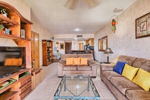 A living room with a TV, sofas, a coffee table, wooden shelves, and a kitchen in the background. - Stunning Views in 2nd Level Condo Princesa B-203 (Puerto Peñasco)