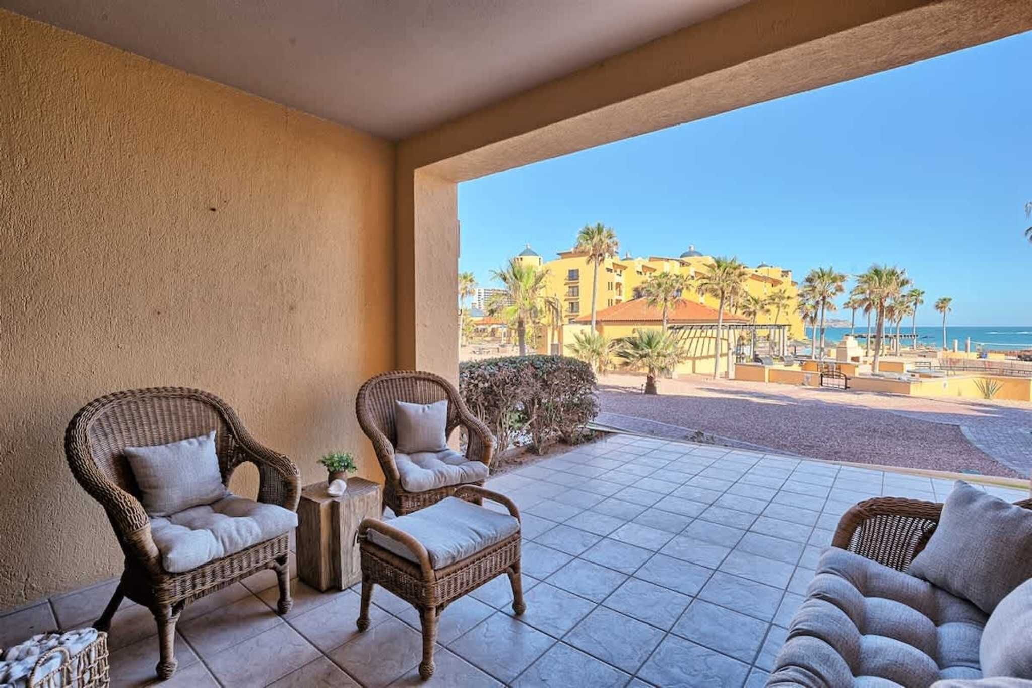 A balcony with wicker furniture, a tiled floor, and a view of a beach and buildings.