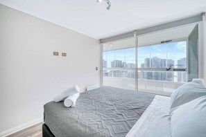 A modern bedroom with a large bed, a view of the city through a sliding glass door, and a cityscape visible through the window.