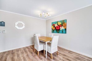 A dining area with a wooden table and white chairs, a wall-mounted painting, a geometric wall decoration, and a ceiling light fixture.