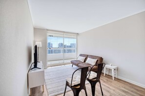 A modern apartment with a large window, a sofa, two chairs, a small table, and a TV on a stand.