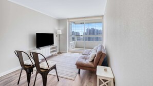Een moderne woonkamer met een flatscreen televisie, twee stoelen, een bank, een kleine tafel en een groot raam met uitzicht op de stad.