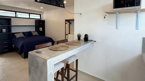 Una habitación pequeña con cama, una mesita con manteles individuales, un televisor montado y un estante para microondas.