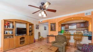 A living room with a television, wooden furniture, a ceiling fan, and a kitchen area with bar stools. - Charming Colorful Condo Near Pool - Pueblos D-101 (Puerto Peñasco)