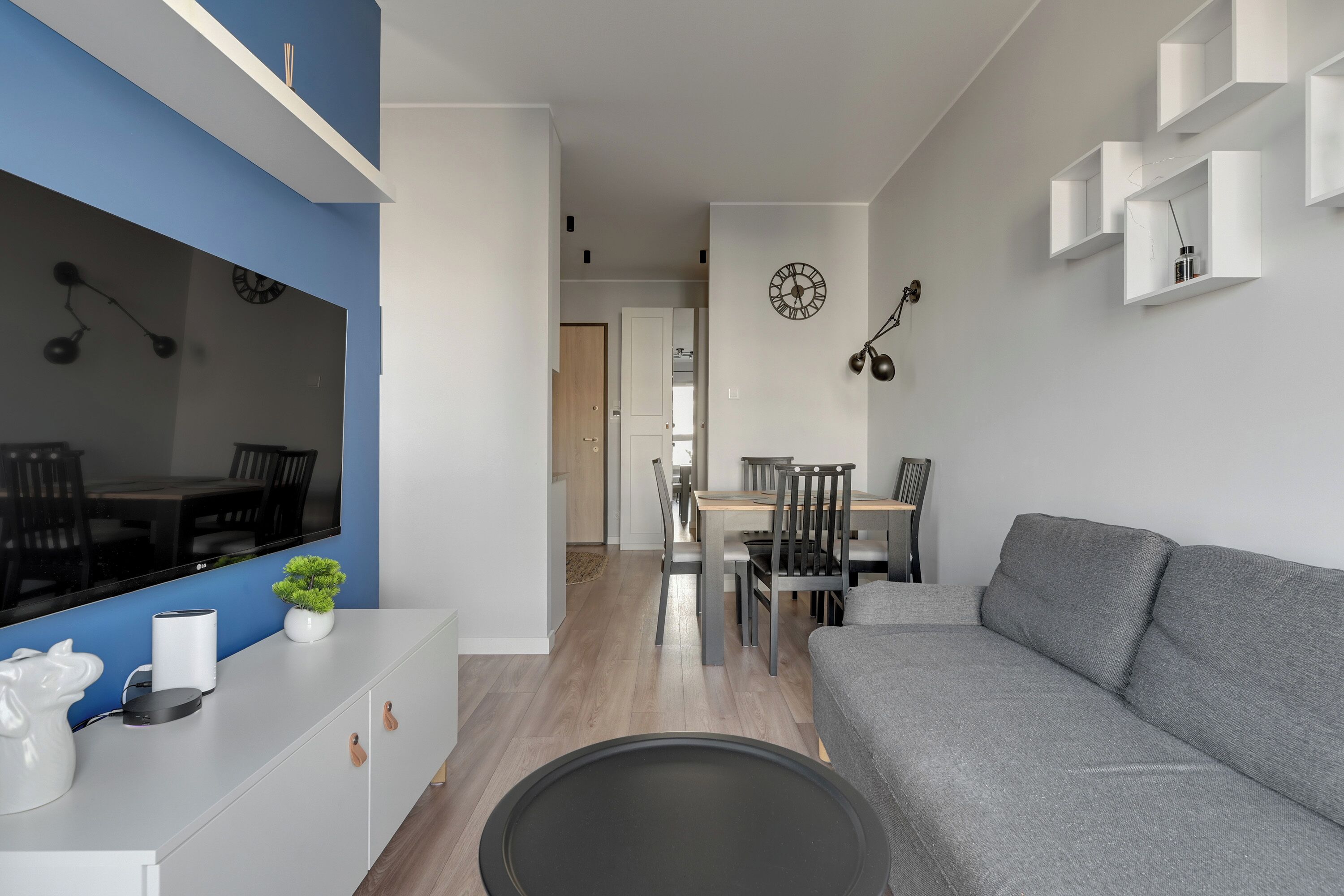 Una sala moderna con un sofá gris, un televisor de pantalla plana, un comedor con sillas y una mesa, y un pasillo que conduce a otra habitación.