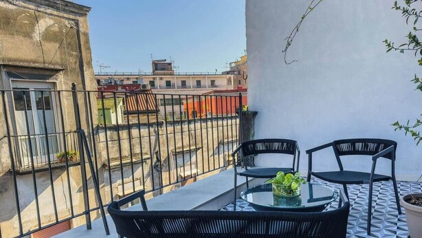 Uma varanda com mesa e cadeiras de vidro, um vaso de planta e vista para prédios vizinhos.