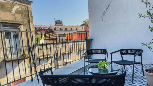 Uma varanda com mesa e cadeiras, um vaso de planta e vista para edifícios vizinhos.
