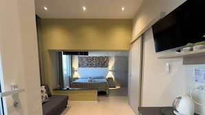 Quarto compacto com cama, mesas de cabeceira, televisão e uma pequena estante com plantas.