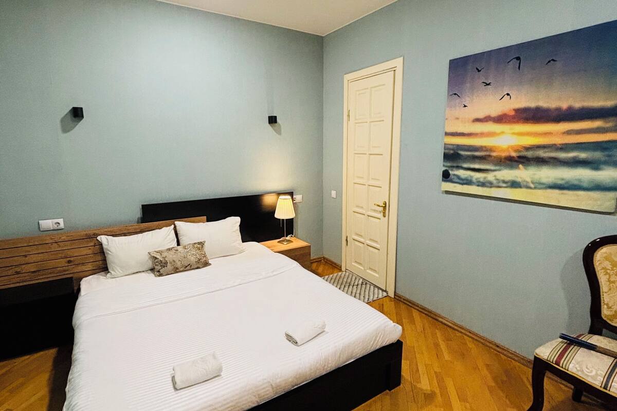 A bedroom with a bed, a nightstand, a chair, a door, and a painting on the wall.