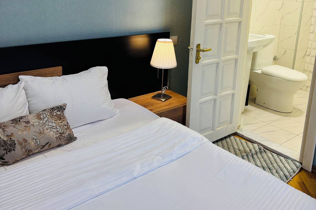 A hotel room with a bed, a nightstand with a lamp, and an open door to a bathroom with a toilet and sink.